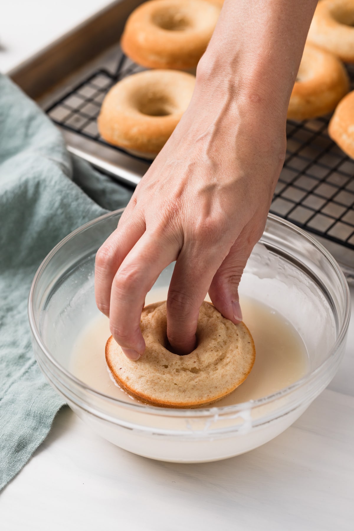 hand dipping baked donut into bowl of glaze with more donuts on wire rack in background