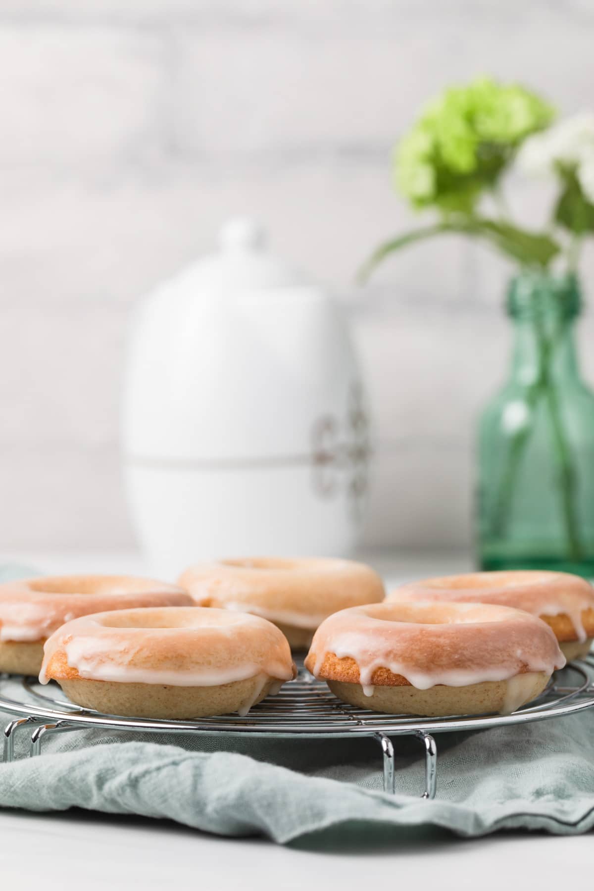 baked donuts arranged in single layer on round metal rack with blue napkin underneath and white tea pot next to glass flower vase