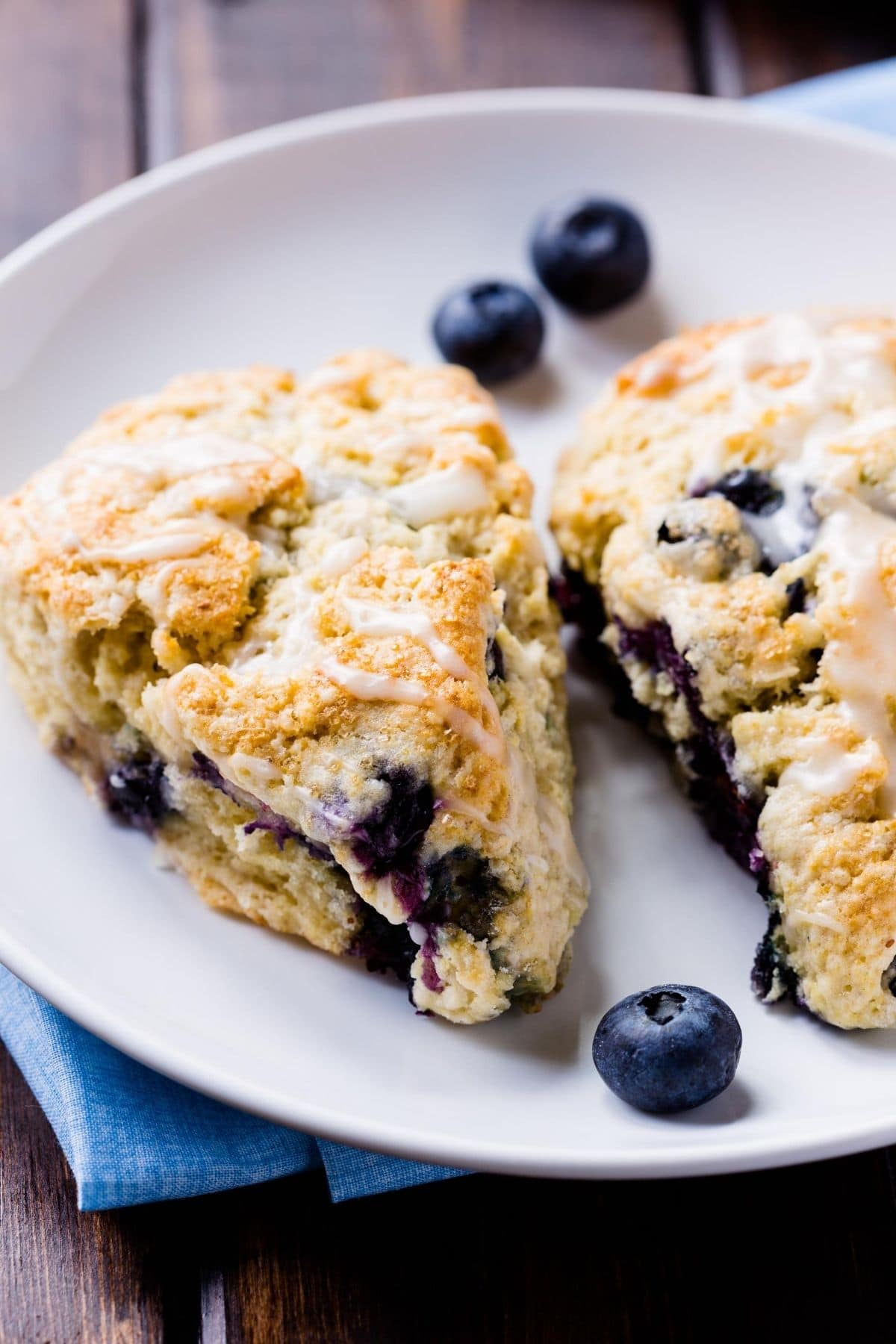two blueberry scones on a white plate with three blueberries set over a blue napkin