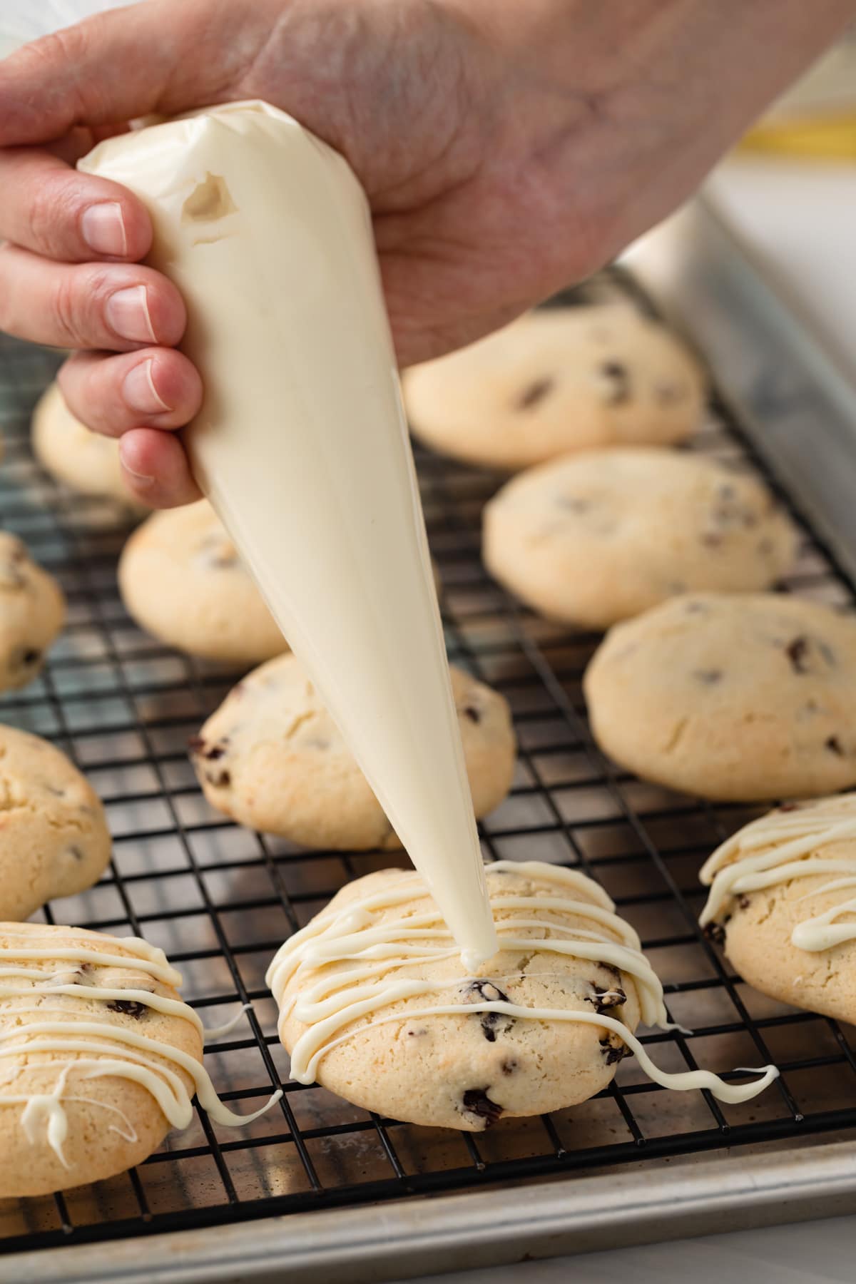 hand using piping bag to drizzle white chocolate over cherry almond cookies on a wire rack.