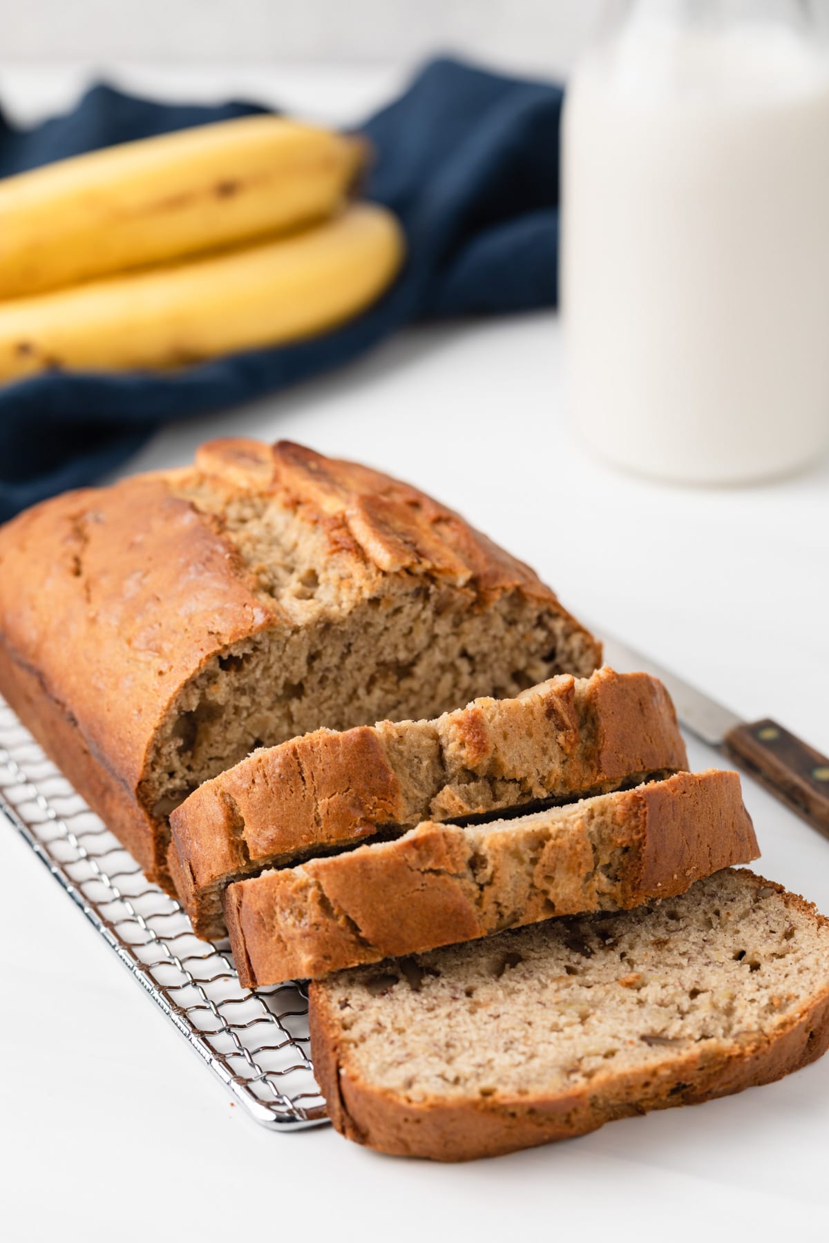 angled view of easy banana bread loaf with three slices cut out and two bananas with jar of milk in background
