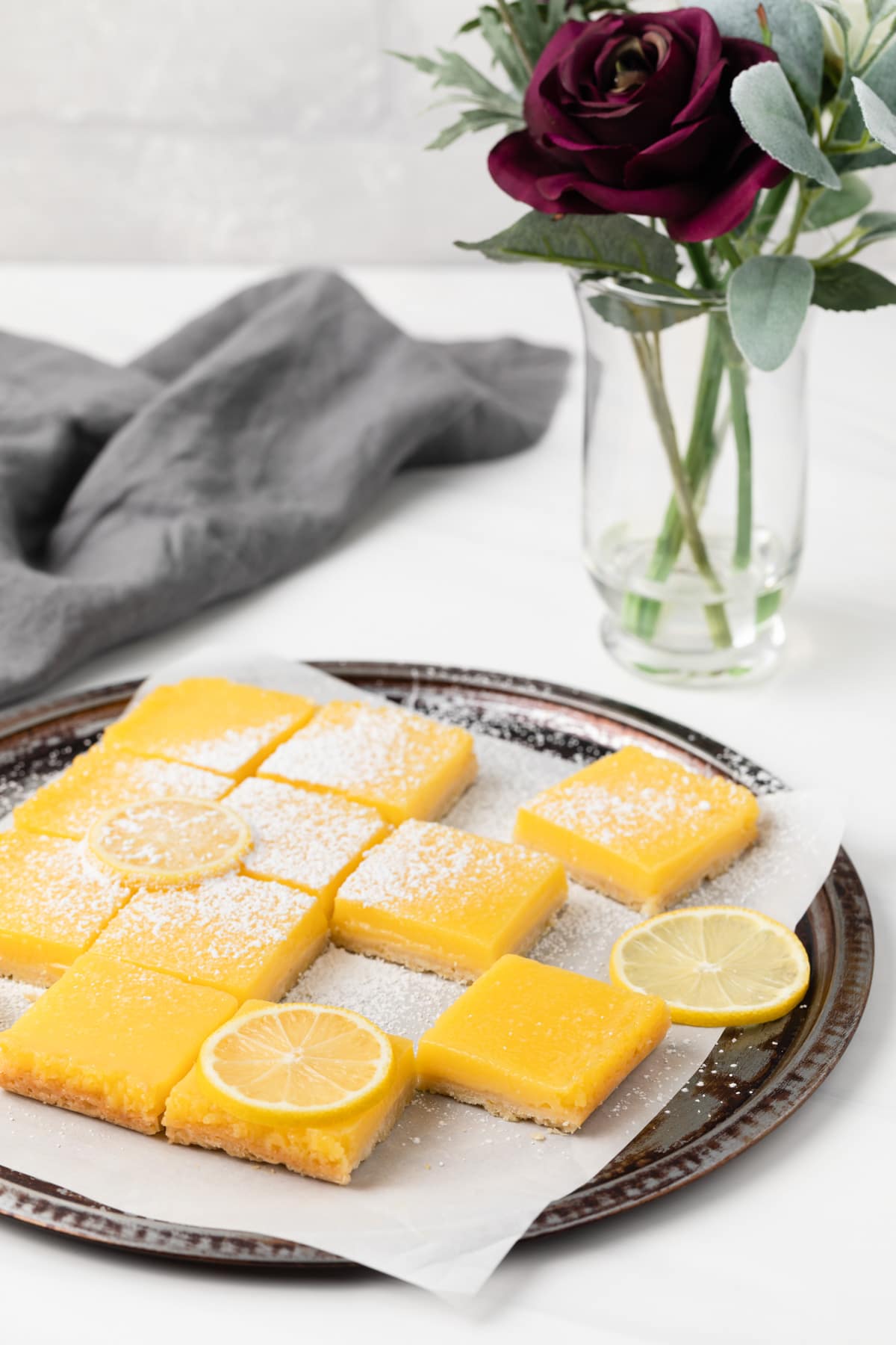 lemon bars arranged on round baking tray lined with parchment paper next to a vase of flowers and grey cloth