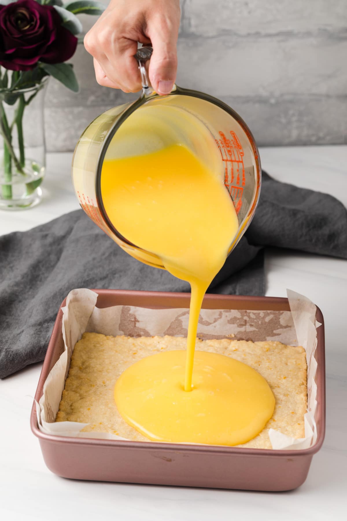 lemon filling being poured over shortbread crust in pink baking pan lined with parchment paper
