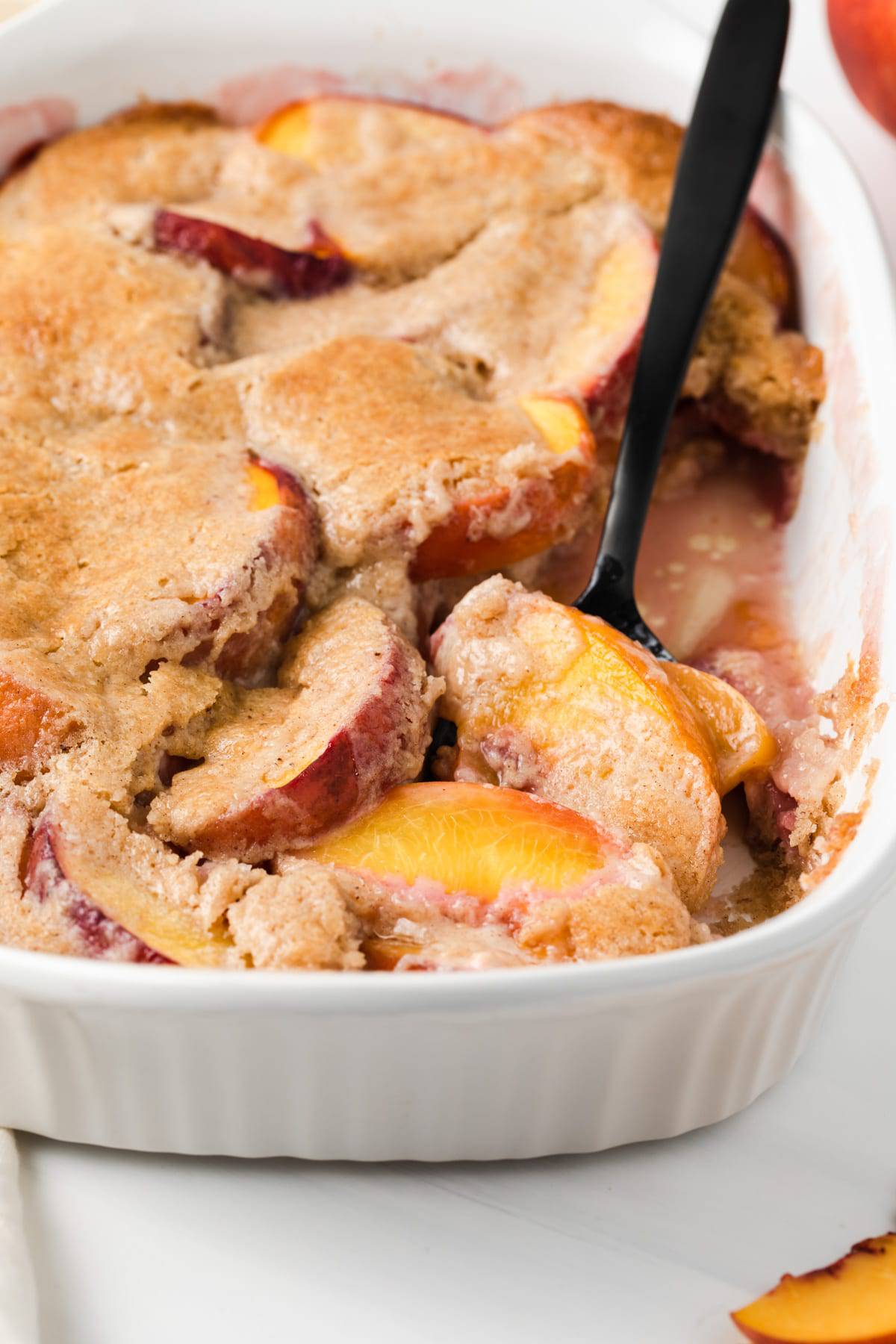 angled view of peach cobbler in white casserole with black spoon