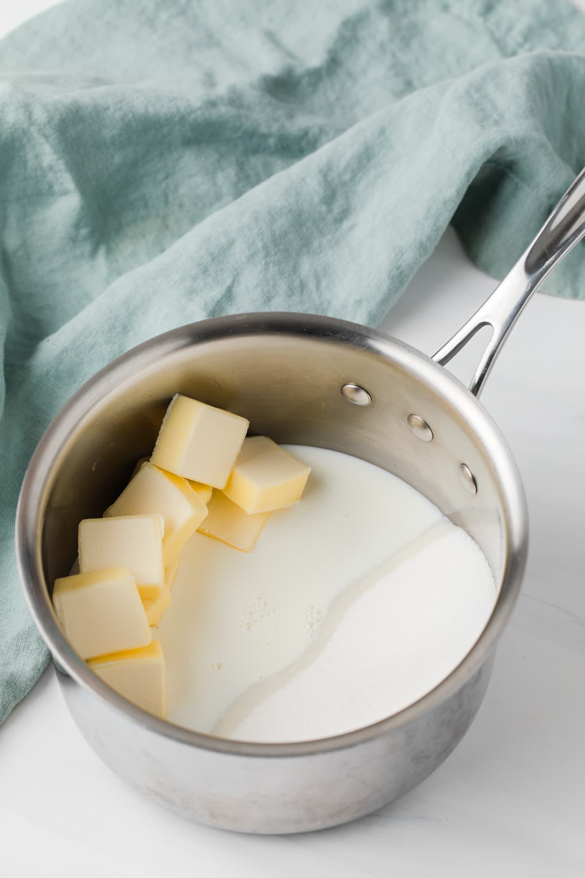 butter, sugar, and milk in stainless steel pot with light blue-green cloth next to it