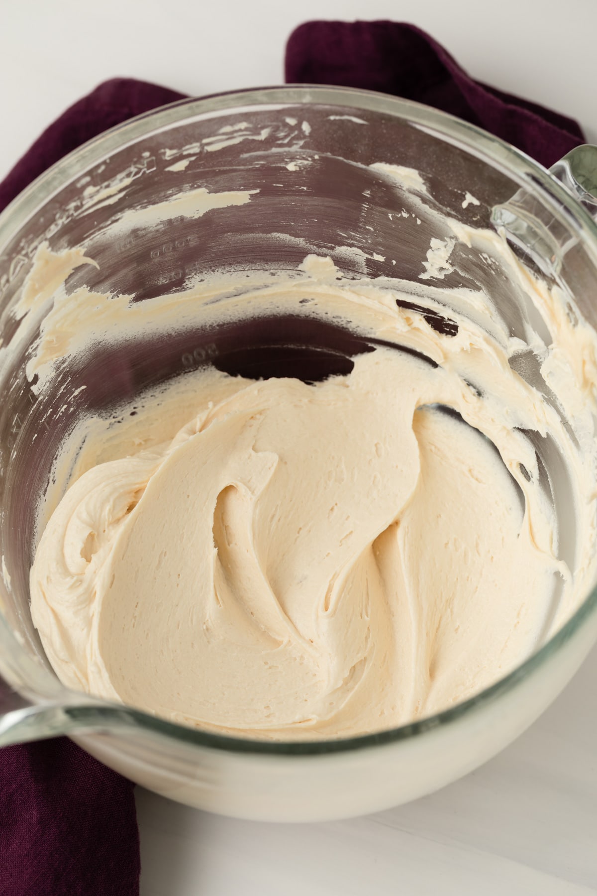 angled view of caramel frosting in large glass mixing bowl with purple cloth next to it