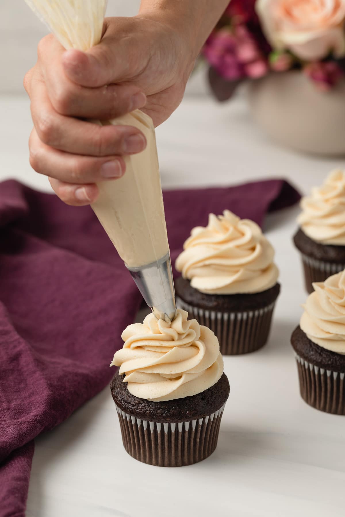 hand piping salted caramel frosting over a chocolate cupcake with purple cloth and more cupcakes in the background