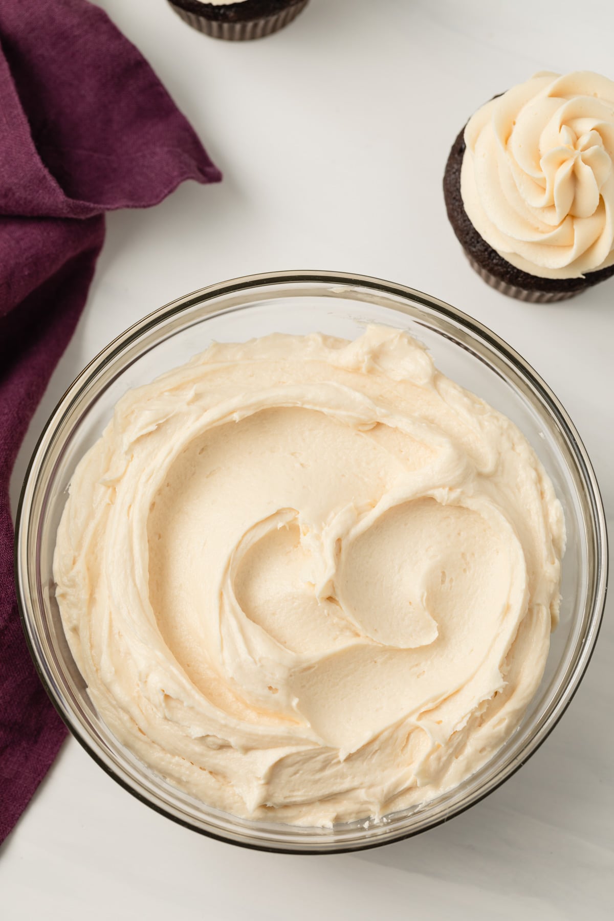 overhead view of salted caramel frosting in glass bowl next to purple cloth and chocolate cupcakes with frosting swirled on top