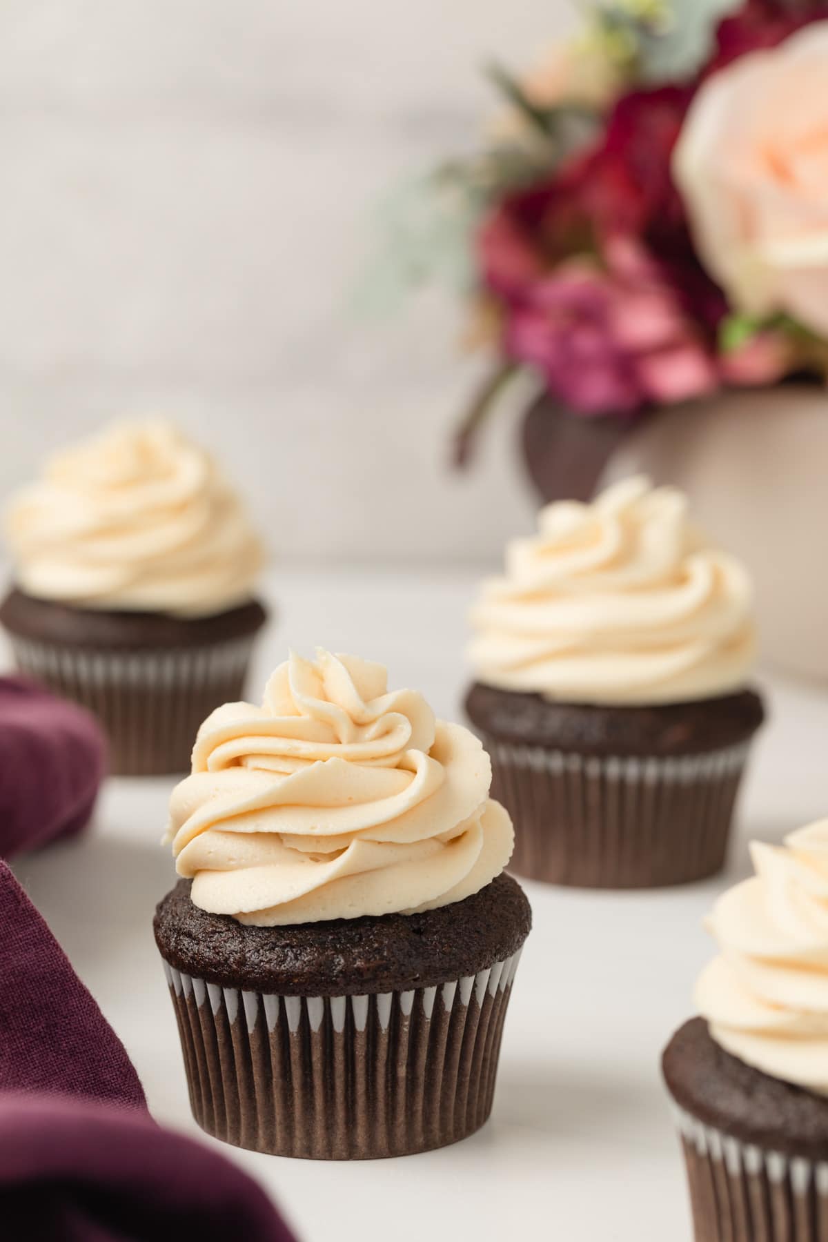 four chocolate cupcakes topped with a swirl of salted caramel frosting with purple cloth to the side and colorful flowers in the background