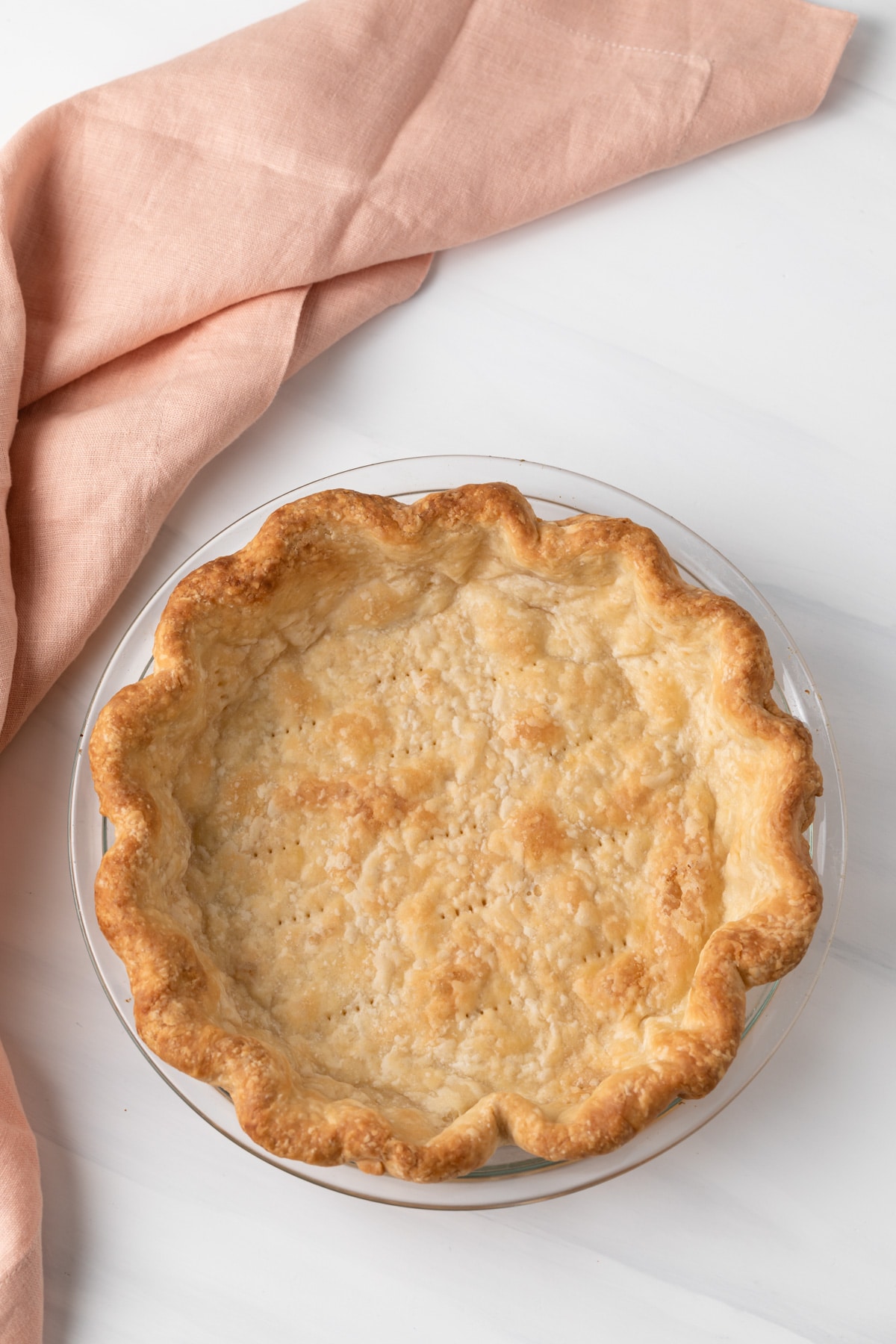 overhead of blind baked pie crust in glass pan with peach colored napkin