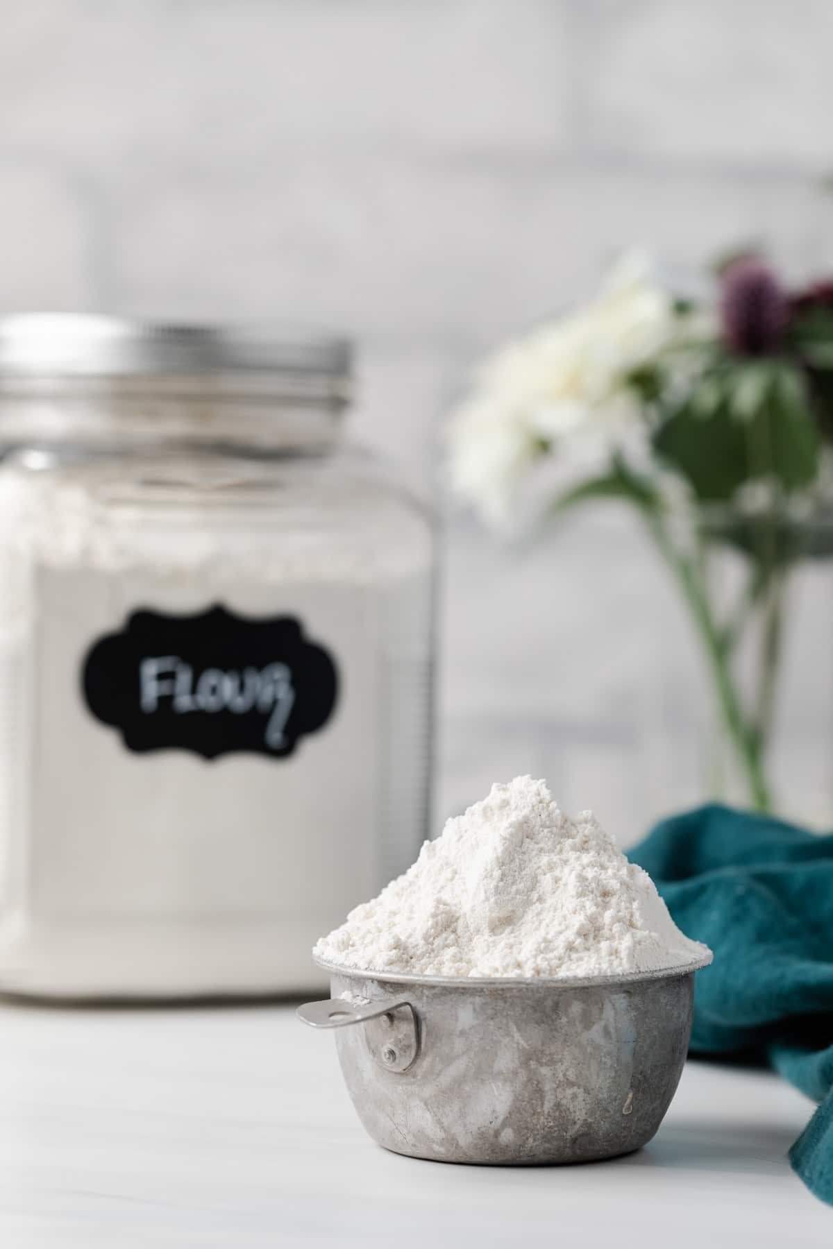 flour piled high in old measuring cup with jar of flour and vase of flowers in background