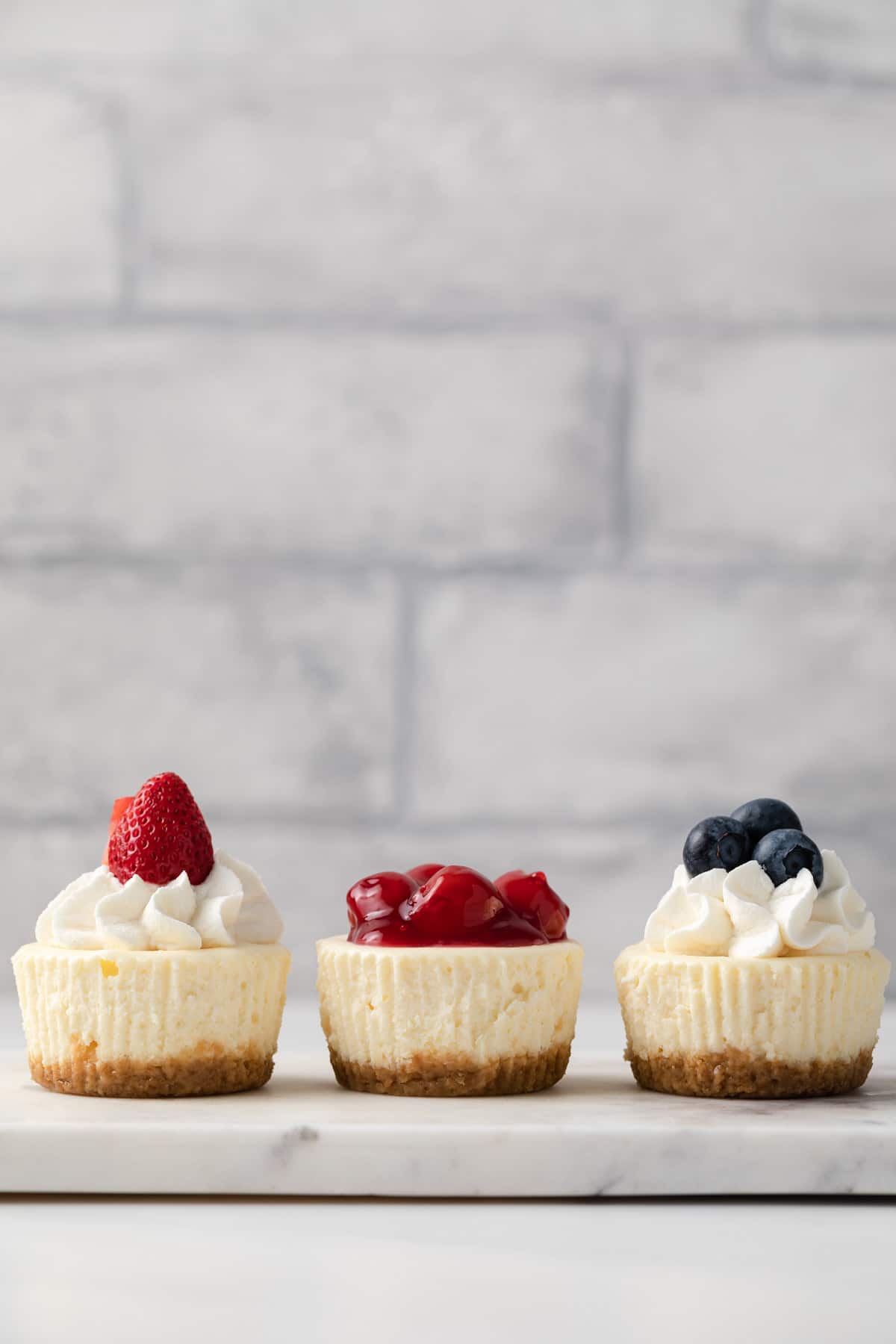 three mini cheesecakes topped with whipped cream and fruit on a marble platter