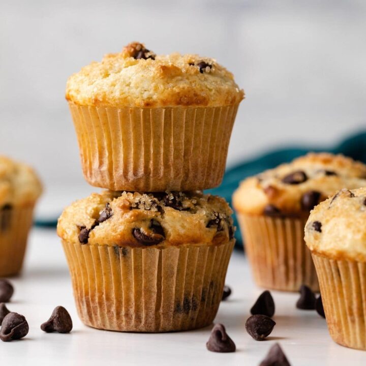 two chocolate chip muffins stacked with more muffins and chocolate chips scattered around