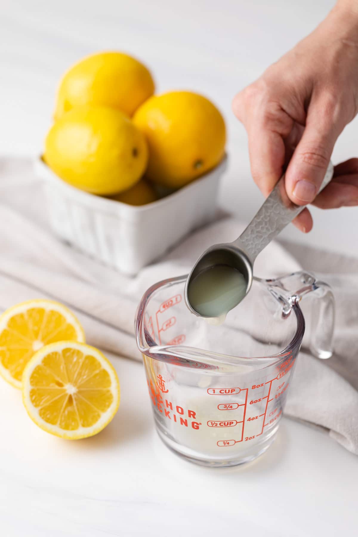 a tablespoon of lemon juice being poured into a glass measuring cup with lemons and napkin in background