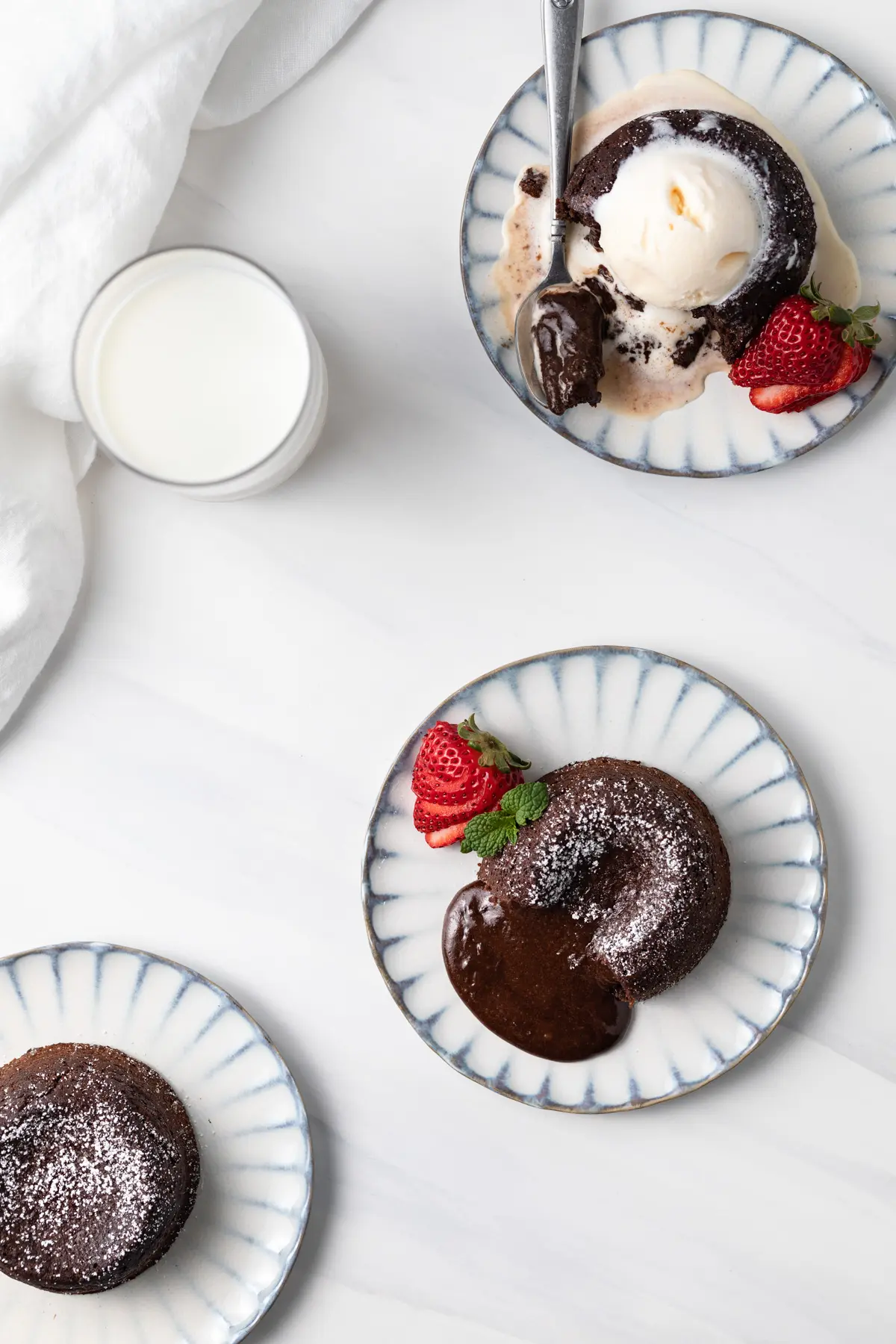 overhead of three individual molten lava cakes with ice cream on one and strawberries, along with a glass of milk