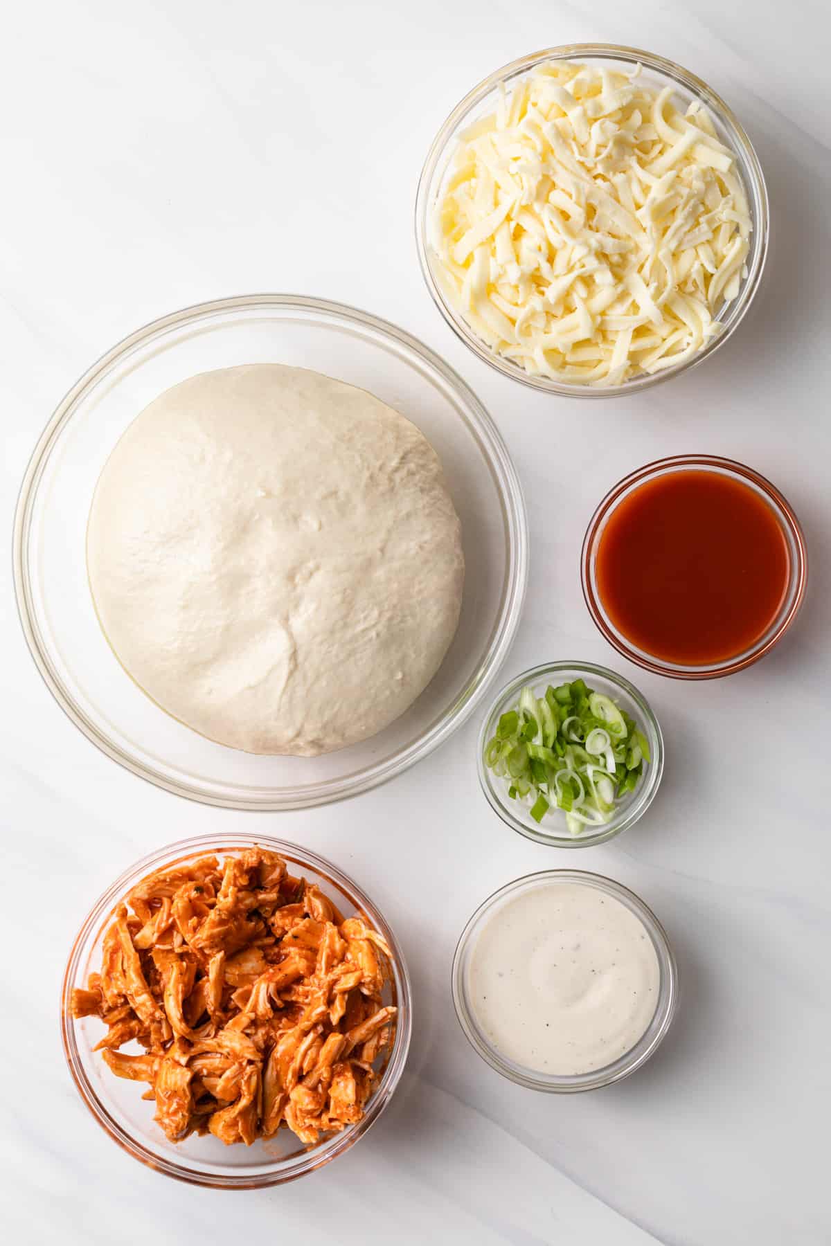 bowl of pizza dough, bowl of mozzarella cheese, bowl of hot sauce, bowl of buffalo chicken, bowl of ranch, and bowl of chopped green onions