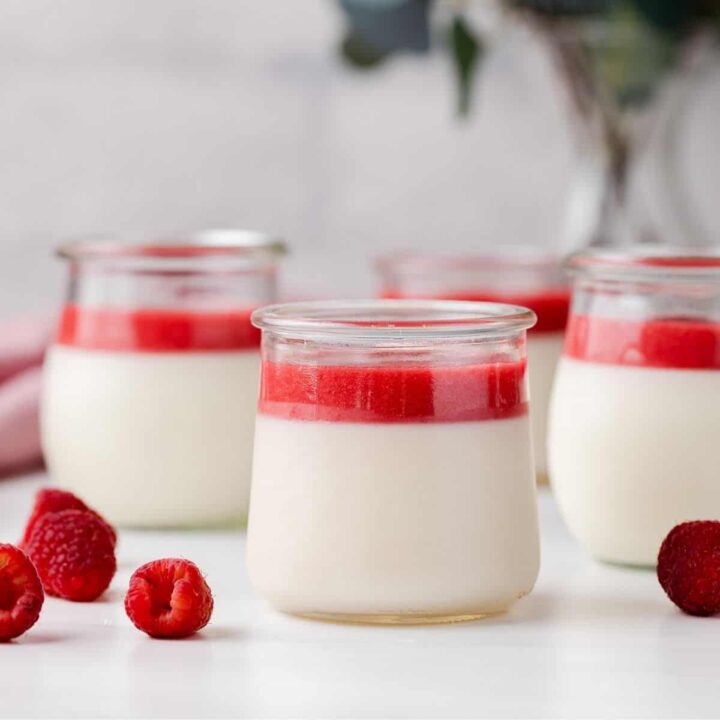 front view of four jars of panna cotta topped with raspberry sauce, fresh raspberries scattered on tabletop and eucalyptus in a vase in the back ground