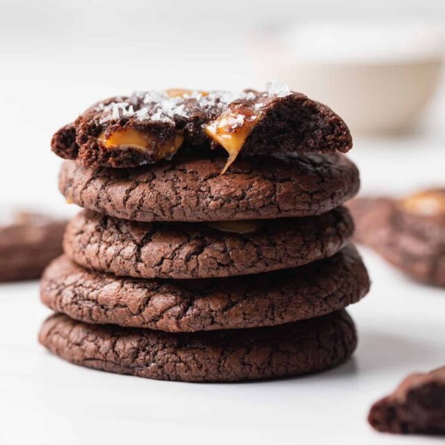 Five salted caramel brownie cookies stacked with the top one broken in half so the gooey caramel is visible.