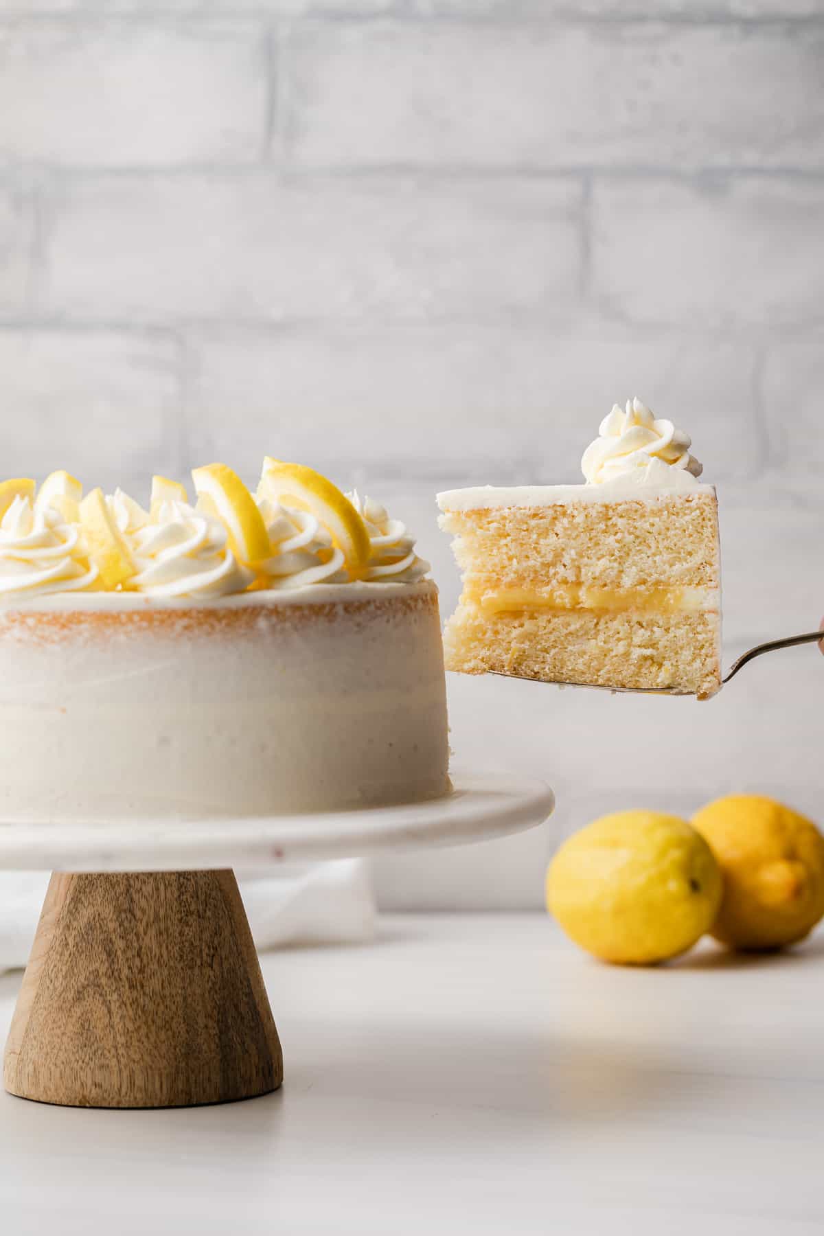 slice being taken out of lemon cake on a cake stand with two lemons in the background