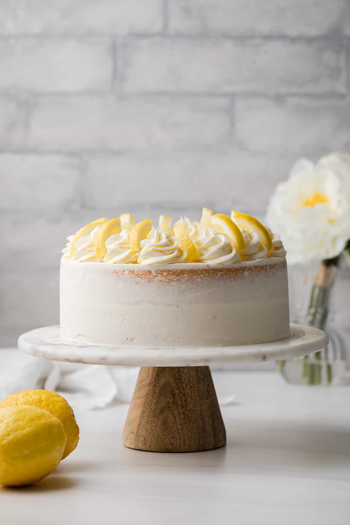 lemon cake on a marble cake stand with wooden bottom, flowers in the background, and l two lemons in the foreground