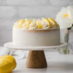 lemon cake on a marble cake stand with wooden bottom, flowers in the background, and l two lemons in the foreground