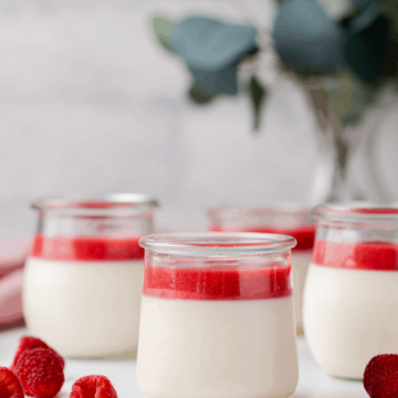front view of four jars of panna cotta topped with raspberry sauce, fresh raspberries scattered on tabletop and eucalyptus in a vase in the back ground