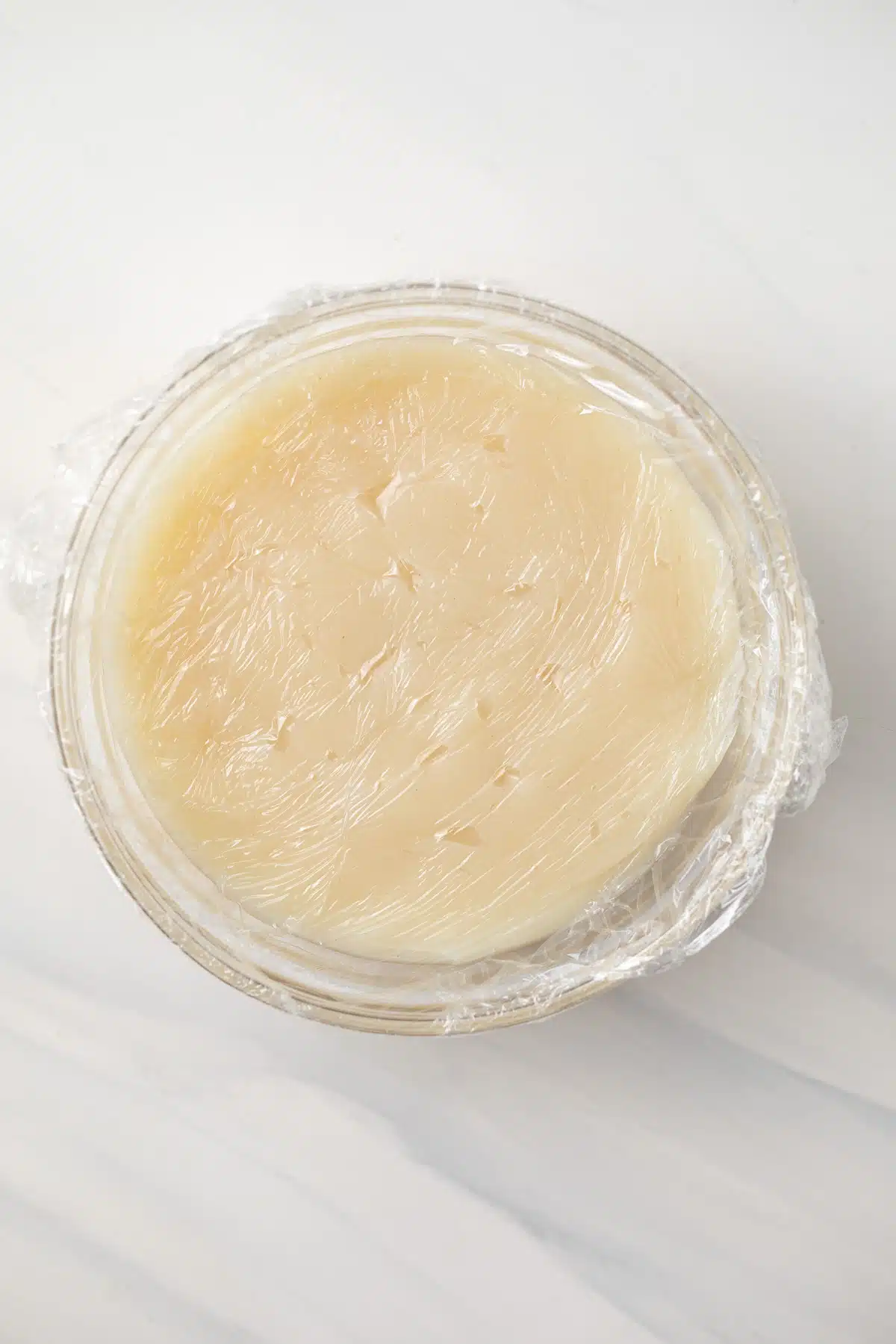 Cooked flour, sugar, and milk in glass bowl with plastic wrap on top.