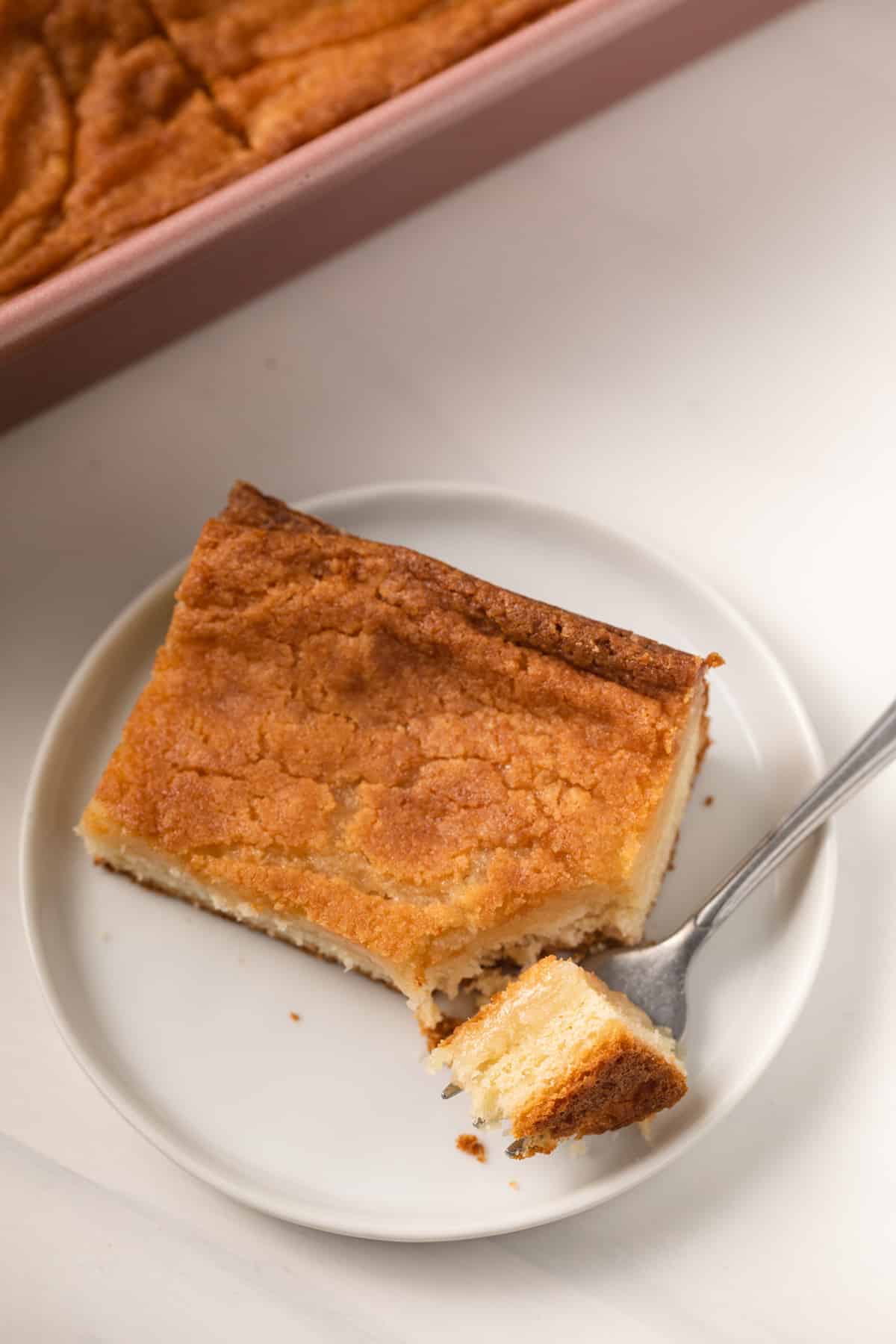 Slice of gooey butter cake on white plate with fork taking a bite out.