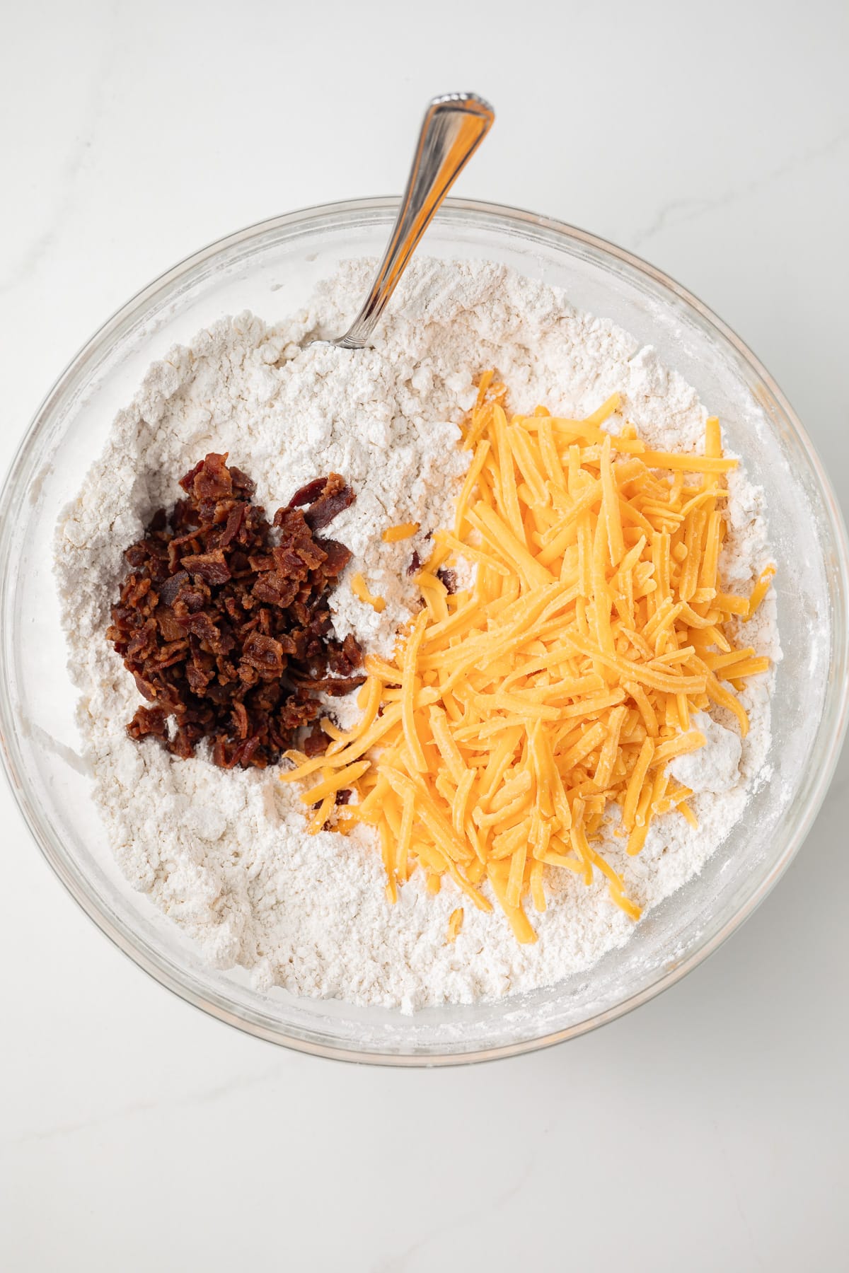 Flour with bacon crumbles and shredded cheddar in a glass bowl.